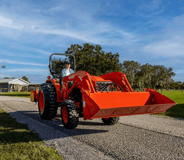 A person operating a Kubota L3302 compact tractor with an LA526 front loader and a rear Land Pride box blade to grade a gravel driveway during spring property maintenance.