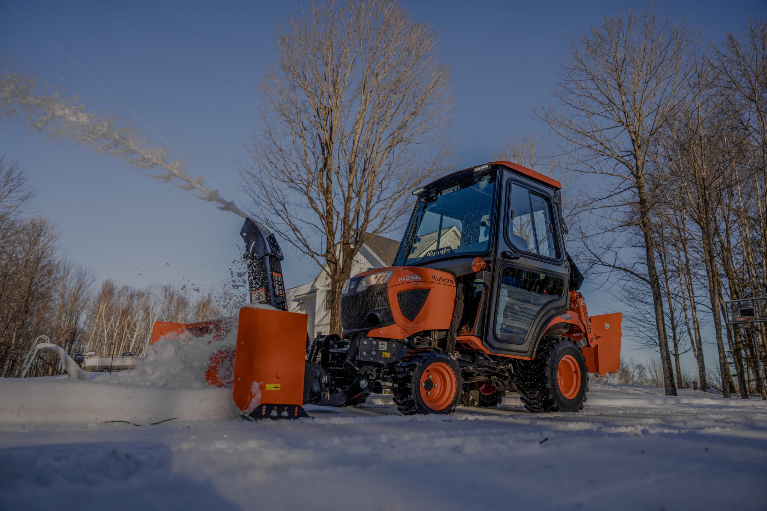 Kubota BX2380 sub-compact tractor with a front-mounted snow blower clearing a snowy driveway. The tractor features an optional Curtis cab, blowing snow into the air on a sunny winter day with trees and a house in the background. Cab shown sold separately.
